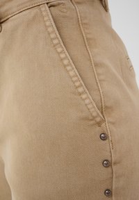 Gros plan sur un tissu en denim beige montrant la couture de la poche avant et trois rivets métalliques sur le côté du pantalon.