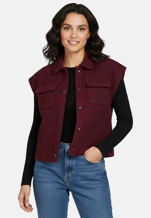 Vrouw met schouderlang donker golvend haar draagt een mouwloos, gestructureerd bordeauxrood vest over een zwarte top met lange mouwen en blauwe spijkerbroek, glimlachend.