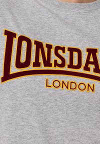 Pilkas medvilninis marškinėliai su dideliu bordo ir geltonu "LONSDALE" tekstu su išlinktu pabrėžimu, o žemiau – "LONDON" atitinkama spalva.