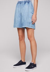 Pantalones cortos de denim azul claro con cintura elástica, que cuentan con un ajuste holgado y dobladillos terminados, acompañados de zapatillas oscuras azul marino.