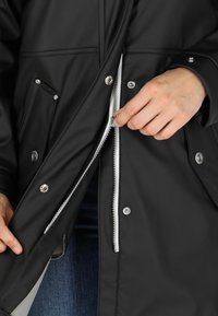 Svart vattentät jacka med dragkedja fram, tryckknappar och sidofickor; slät textur med ett varmt foder synligt vid öppningen.