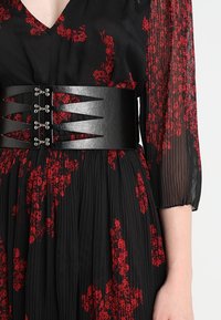 Ceinture en similicuir noir avec découpes angulaires et ferrures métalliques, portée sur une robe plissée à motif floral rouge.