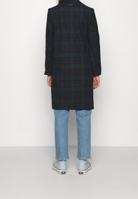 Kostkovaný kabát v tmavě námořnické modři a zelené barvě, s dlouhým rukávem a přímým střihem. Nositelný přes světle modré džíny a modré boty s černými doplňky.