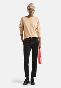 Frau trägt einen beigen Pullover, schwarze Hose, schwarze Schuhe, eine mehrfarbige gestreifte Mütze und hält eine kleine rote Handtasche.