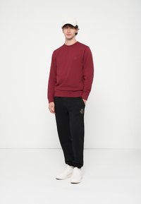 Bordó pulóver, fekete melegítőnadrág arany hímzéssel, fehér tornacipő. Egyszerű kialakítás, testhez simuló szabás, pamut anyag. Modell egyszínű háttér előtt áll.