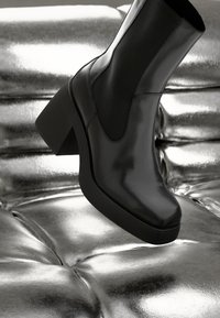 Black ankle boot with a glossy leather finish, square toe, elastic side panels, and a chunky heel, set against a shiny metallic background.