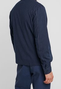 Camisa de rayas azul marino con mangas largas, con rayas verticales blancas, detalle de puño con botones y un diseño limpio y aerodinámico.