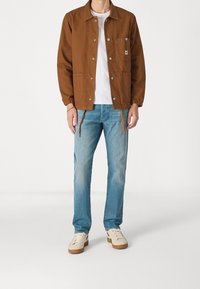Brun bomullskavaj med tryckknappar, två framfickor och dragsko i nederkant, tillsammans med ljusblå denimjeans och beige skor.