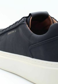 Sneaker nera con materiale liscio, suola in gomma bianca a contrasto, dettagli in pelle e lacci texturizzati. Presenta il marchio "SMITH X".
