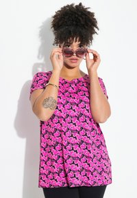 Top de manga corta rosa con un patrón de corazones en negro, con un corte holgado y un escote redondo. El modelo lleva gafas de sol y accesorios mínimos.