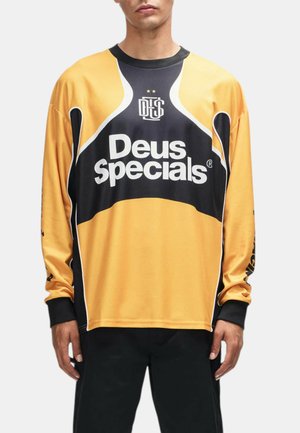 Persona che indossa una maglietta gialla e nera a maniche lunghe con il testo e il logo "Deus Specials" sulla parte anteriore, abbinata a pantaloni neri.