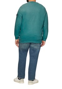 Sweatshirt teal avec des poignets et un ourlet côtelés, associé à un jean bleu délavé et des baskets blanches, montrant une vue arrière décontractée.