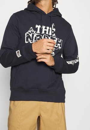Mann trägt dunkelblauen The North Face Hoodie mit weißem Logo und beige Hose, Hände vor der Brust verschränkt, vor schlichter hellem Hintergrund.