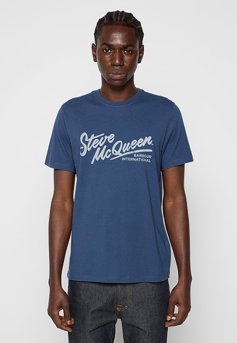 Camiseta de algodón azul marino con un estampado gráfico en blanco que incluye el texto "Steve McQueen" y "Barbour International". Tiene mangas cortas y un cuello redondo.