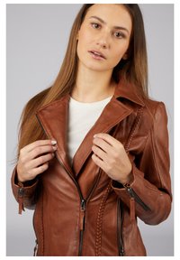 Braune Lederjacke mit einem spitzen Kragen, asymmetrischem Reißverschluss und geflochtenen Details an der Vorderseite. Das Model trägt ein weißes Oberteil darunter.