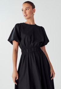 Črna obleka s kratkimi napihnjenimi rokavi, elastičnim pasom in tekočo krilo. Narejena iz gladkega materiala, ima okroglo izrez in preprost dizajn.