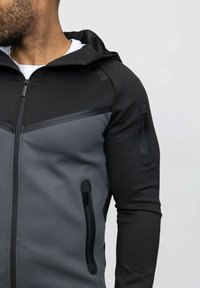 Veste zippée noire et grise avec capuche, présentant un design géométrique, un tissu texturé et des poches latérales zippées.