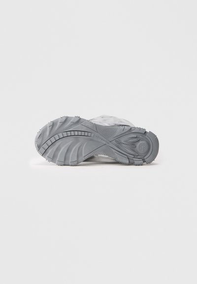 Semelle extérieure de chaussures de sport gris clair avec un caoutchouc texturé, des rainures profondes et de petites crampons pour une meilleure adhérence et stabilité.
