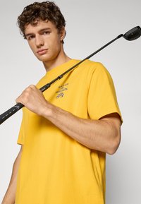 Rumena bombažna majica s krožnim izrezom, na kateri je v modri barvi natisnjen napis "le CLUB du MALBÓN". Na rami leži golf palica.