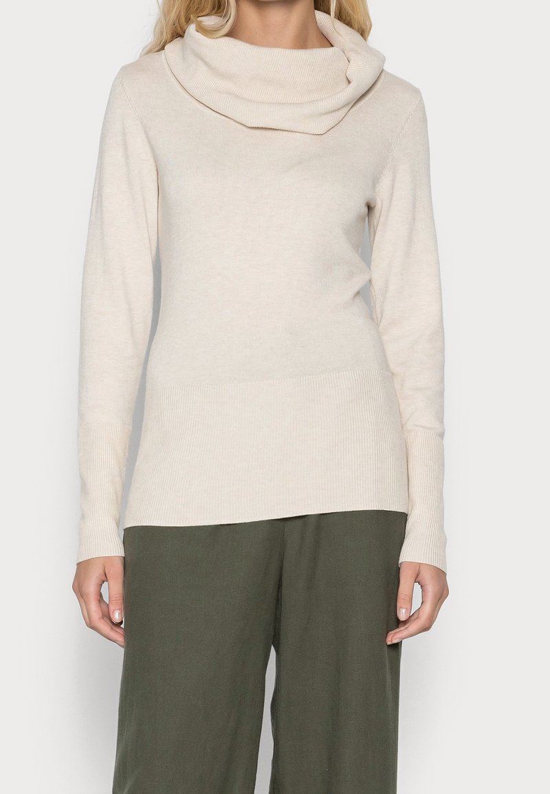 Beige Strickpullover mit Kaskadenkragen, langen Ärmeln, gerippten Bündchen und einem etwas längeren hinteren Saum, kombiniert mit loser grüner Hose.