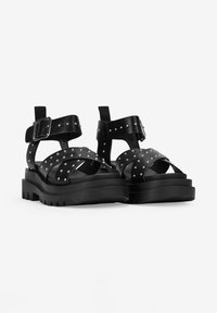 Sandalias de cuero negro con suela gruesa, detalles de tachuelas, hebilla ajustable y tiras cruzadas. Diseño texturizado y duradero.