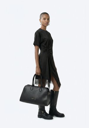 Femme portant une robe noire à manches courtes avec un ourlet en dentelle, des bottes noires montantes jusqu'aux genoux, tenant un grand sac à main noir Valentino.