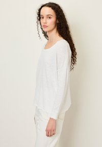 Noor naine, kellel on pikk lokkis juuksed, seisab küljepealt, seljas lahtine valge pikkade varrukatega särk ja valged püksid neutraalse tausta ees.