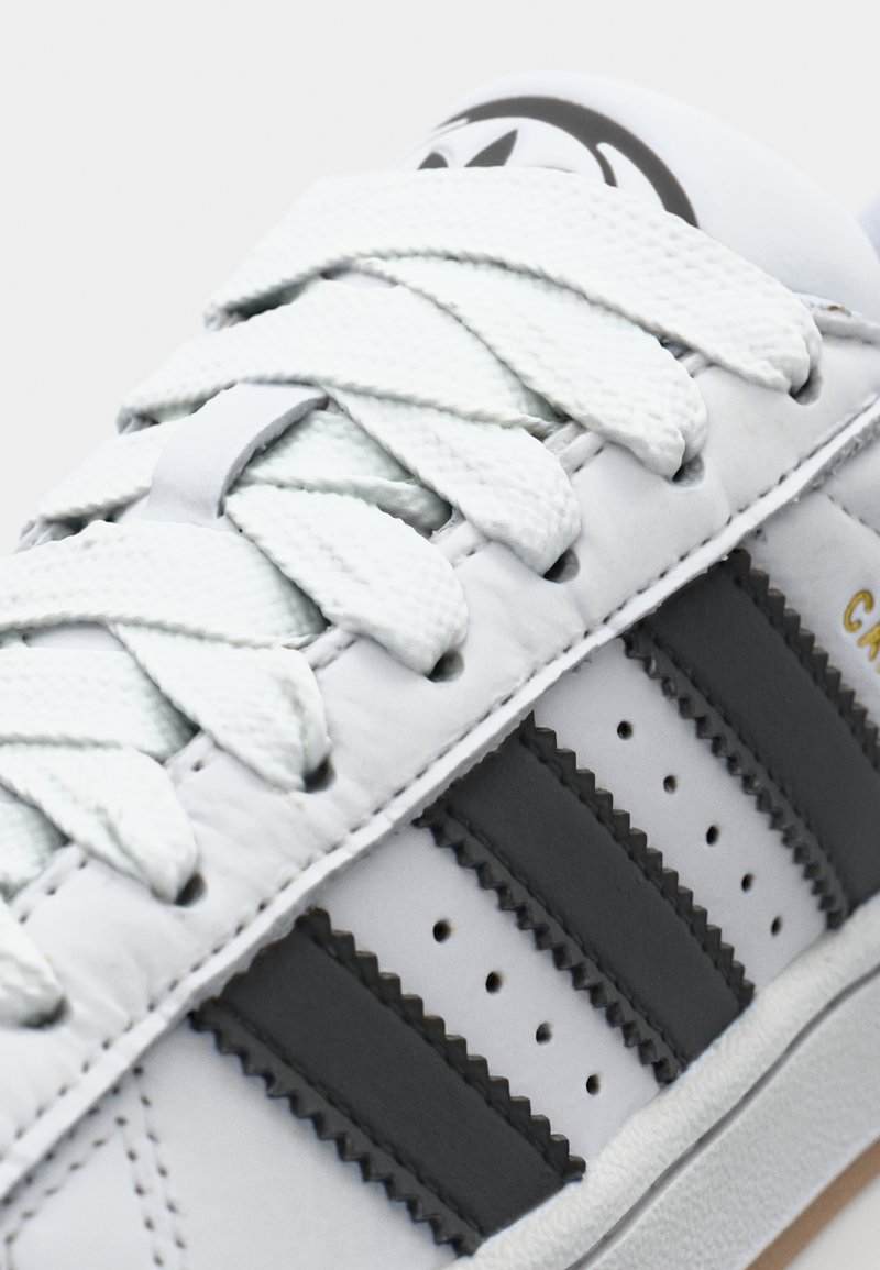 Zapatilla de cuero blanco con rayas grises, cordones texturizados y detalles de diseño perforados. Presenta una puntera redonda y suela de goma.