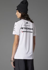 Camiseta atlética blanca de manga corta, con logotipos negros que incluyen "PETRONAS" y "CROWDSTRIKE". Usada con un gorro negro.