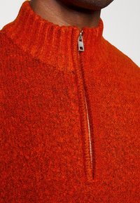 Roter Wollpullover mit hohem Kragen und kurzem Reißverschluss vorne. Die Textur ist dick und strukturiert, zeigt ein weiches, gestricktes Muster.
