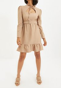 Beiges langärmliges Kleid mit rüschenförmigem Saum, quadratischem Ausschnitt, frontaler Öffnung und einem Gürtel. Kombiniert mit geflochtenen beigen Riemchensandalen.