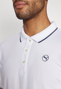 Biely polo tričko s námorníckym golierom, tromi gombíkmi a kruhovým logom s vyskočenou mačkovitou šelmou. Hladká textúra látky.