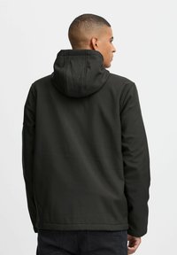 Veste à capuche noire avec une texture lisse, dotée d'un détail de couture sur la capuche et d'une coupe droite à l'ourlet, portée sur un pantalon sombre.