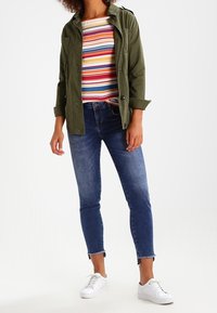 Olijfgroene jas, gestreept overhemd met lange mouwen met rode, blauwe en gele strepen, blauwe skinny jeans, witte sneakers. Casual outfit met verschillende texturen.