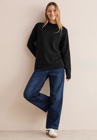 Maglione nero a maglia con design a coste sulle spalle, abbinato a jeans blu larghi e sneakers bianche. Texture liscia, vestibilità casual.