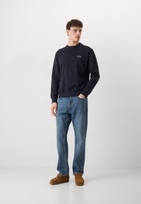 Sudadera azul marino con cuello redondo y logo bordado, combinada con jeans azul claro y zapatos de ante marrón tipo slip-on.