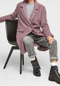 Cappotto lungo in misto lana rosa, con chiusura a un solo petto e due bottoni, indossato sopra un maglione grigio a maglia e jeans grigi, abbinato a stivali neri con lacci.