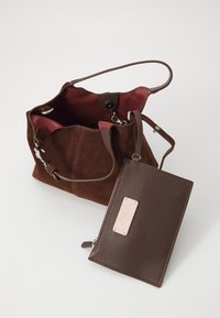Geantă tote din suede maro, cu un interior roșu, dotată cu accesorii din metal argintiu și un portofel detasabil din piele maro, cu un etichetă roz.