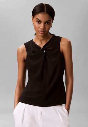 Sima sötét hajú nő ujjatlan fekete felsőt visel, mely kulcslyuk csavart részlettel rendelkezik, fehér pliszírozott nadrággal párosítva, szürke háttér előtt.