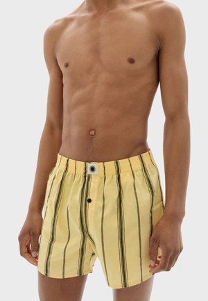 Oben ohne Person in gelben Shorts mit vertikalen schwarzen Streifen, stehend vor einem einfachen hellen Hintergrund.
