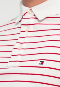 Λευκό πόλο μπλουζάκι με κόκκινες ρίγες, με κλασικό γιακά και τρία κουμπιά, καθώς και μικρό λογότυπο στο αριστερό στήθος. Μαλακό, υφασμάτινο με υφή.