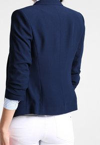 Persona vistiendo un blazer ajustado azul marino con mangas arrugadas sobre una camisa a rayas azul y blanca, combinado con pantalones blancos, de espaldas.