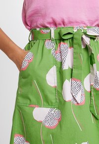 Jupe verte avec un motif floral en rose et noir. Dotée d'une ceinture à nouer, de poches latérales et d'une texture de tissu lisse et léger.