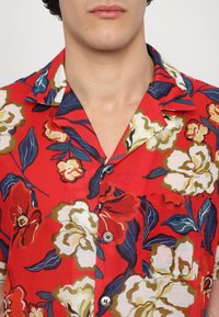 Punainen lyhythihainen paita, jossa on valkoisia, keltaisia ja vihreitä kukkakuoseja; sisältää napitetun etuosan ja rintataskun.