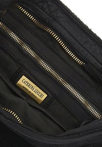 Interno di una borsa a tracolla in pelle nera con più scomparti con zip, foderatura in tessuto nero e un'etichetta in metallo dorato con la scritta "CATERINA LUCCHI."