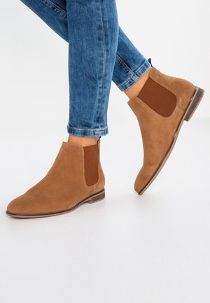Personne portant un jean bleu ajusté et des bottines Chelsea en daim beige à panneaux latéraux élastiques, debout sur une surface blanche.