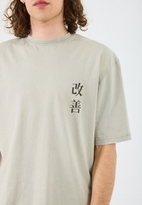 Helles graues T-Shirt aus Baumwolle mit kurzen Ärmeln. Enthält schwarze japanische Zeichen auf der linken Brust. Glatte Textur, klassischer Rundhalsausschnitt.