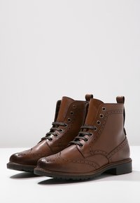 Brune læder snøreankelstøvler med brogue-detaljer, afrundede tæer og en lav hæl. Har metaløjer og en pull tab på bagsiden.
