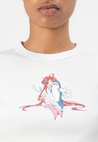 T-shirt blanc présentant un personnage de dessin animé avec des cheveux roux, des accents bleus et des chaussures roses. Tissu lisse avec un design imprimé graphique.