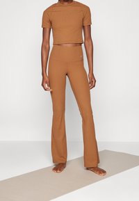 Person standing barefoot on a light yoga mat wearing brown ribbed high-waist flared leggings and a matching short-sleeve crop top.